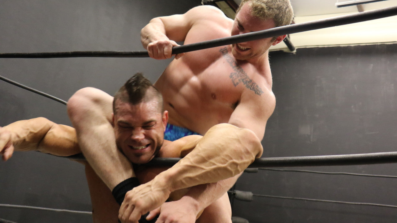 Talon chokes Brian Cage on the ropes with his leg huge pecs